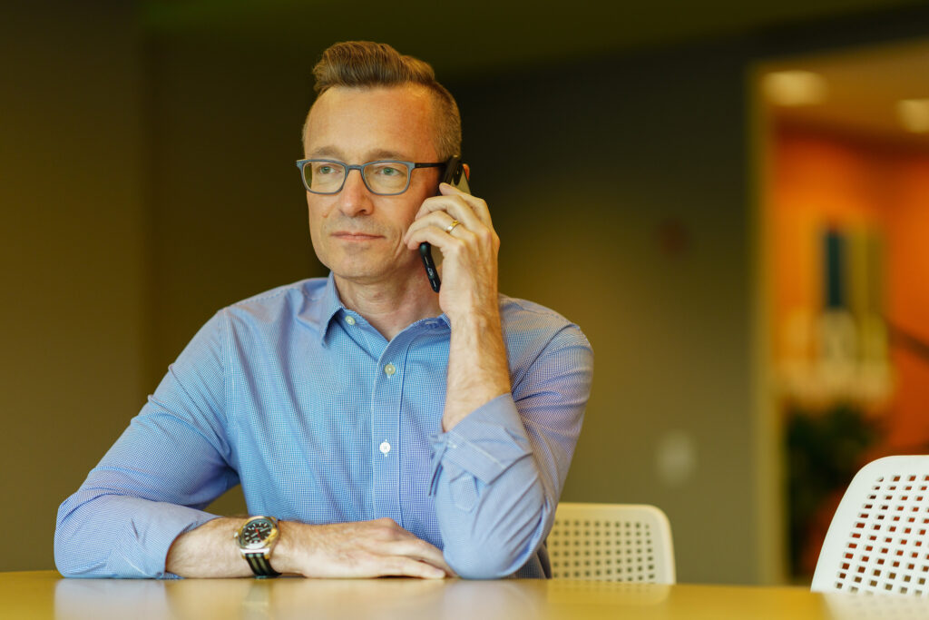 Derek Rockelman in a blue shirt takes a phone call at a table in the bright open office.