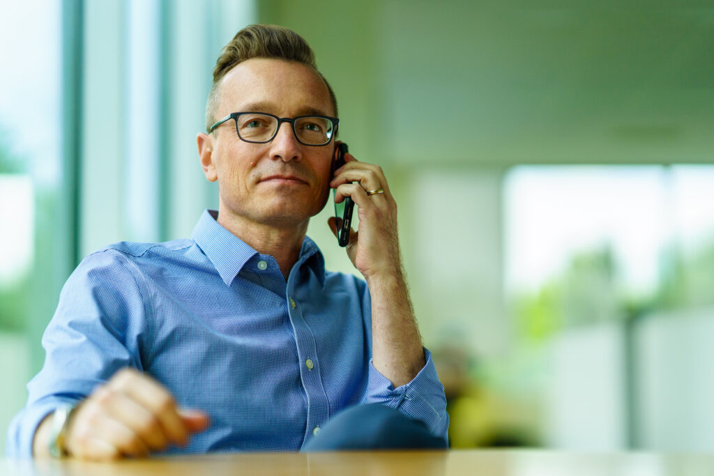 Derek Rockelman in glasses listens on a phone call by the office windows, greenery blurred softly behind him.