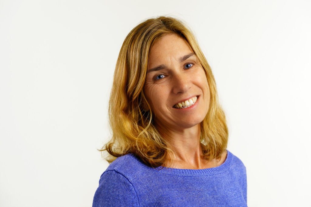 Studio portrait of Wendy Fox smiling in a violet sweater against white, hair falling loose past her shoulders.