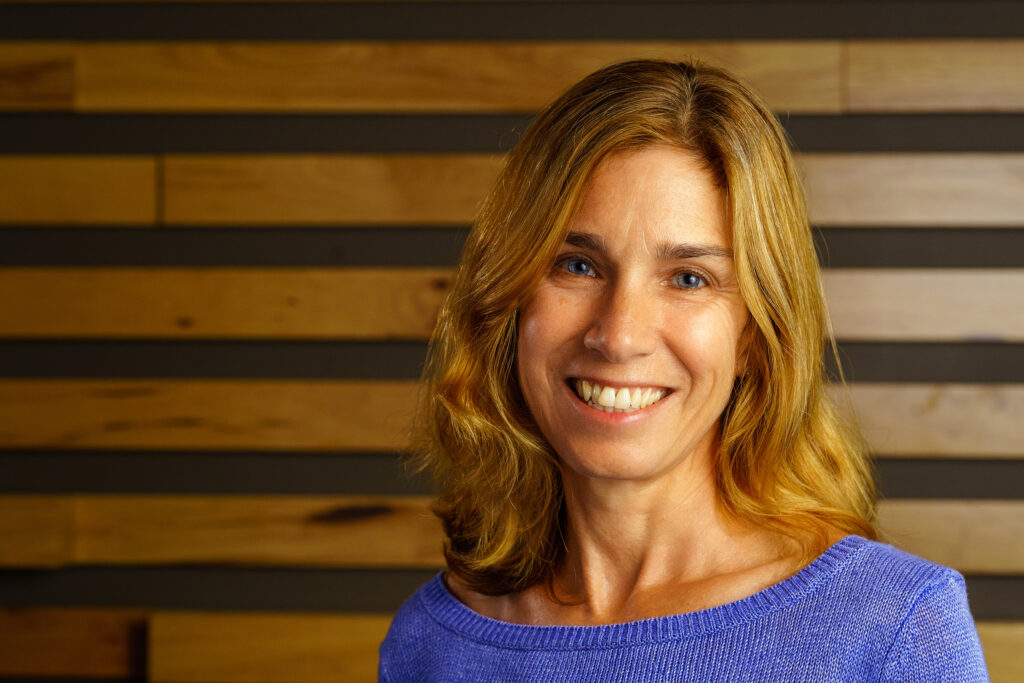 Wendy Fox smiles before a slatted wood accent wall, her violet sweater warm against the striped grain.