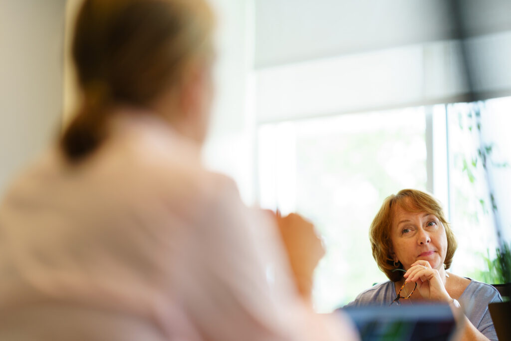 Colleague listens thoughtfully across the table, chin on hand, while a teammate speaks out of focus in the foreground.