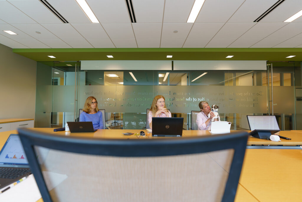 Wide view of three team members on laptops and tablets along the conference table in the green-walled meeting room.
