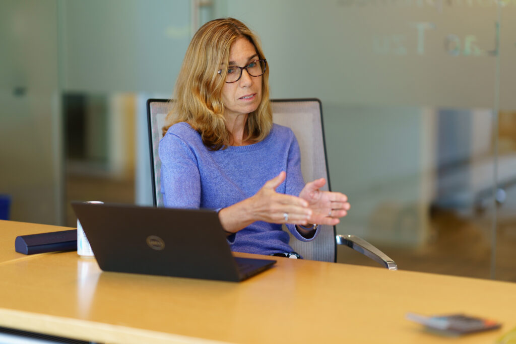 Wendy Fox gestures mid-explanation beside her open laptop, the etched glass wall lettering blurred across the foreground.