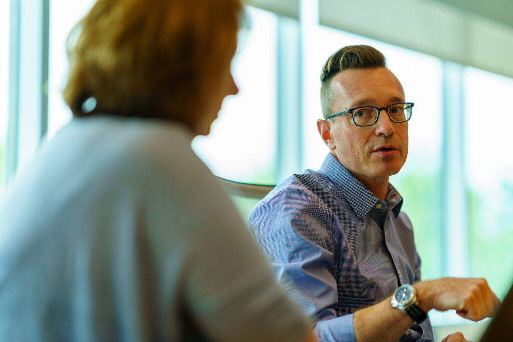 Two colleagues mid-conversation at the conference table, one gesturing in a checked shirt by the window wall.