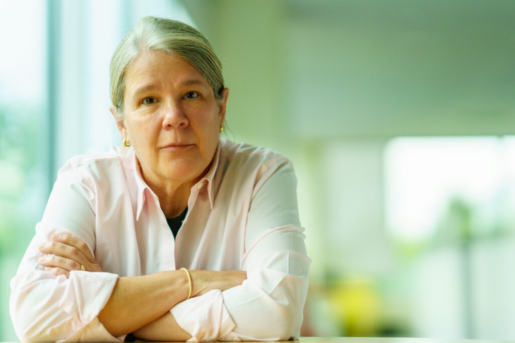 Gina Vanderloop leans on folded arms at the table, soft office light and greenery blurred behind her.