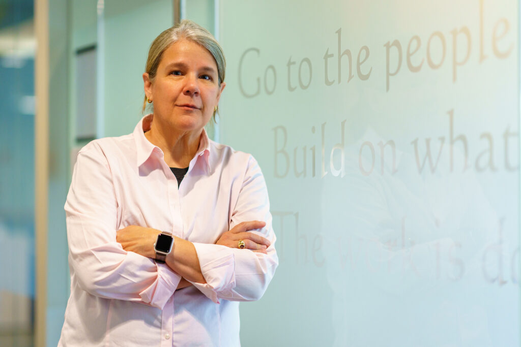 Gina Vanderloop stands with crossed arms before an office wall inscribed Go to the People, Build on What They Have.