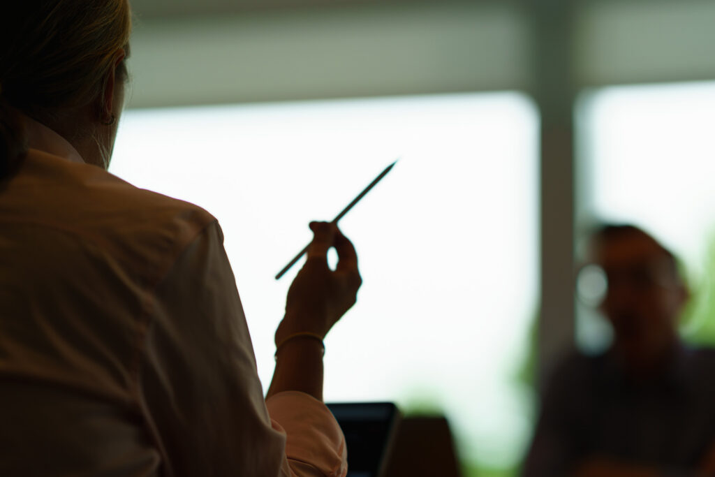 Silhouetted colleague raises a pencil in the foreground while the meeting continues across the sunlit conference room.