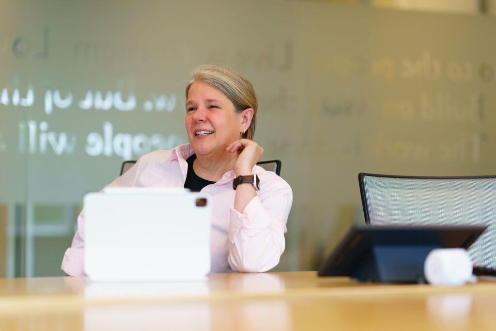 Gina Vanderloop laughs at the conference table beside a tablet, mission lettering etched across the glass behind her.