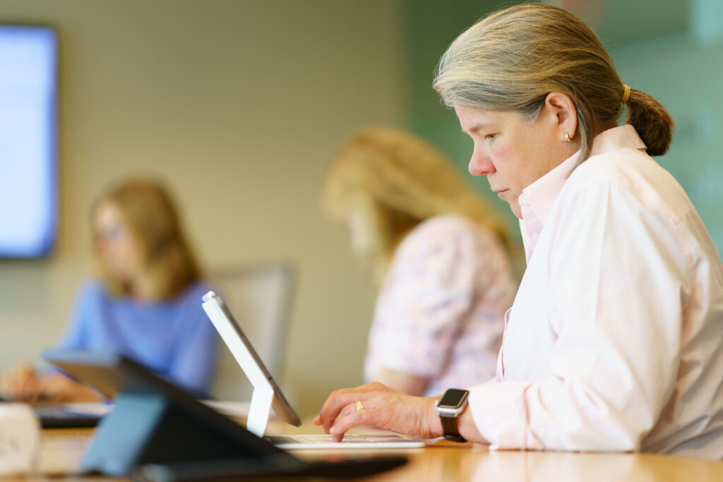 Gina Vanderloop types on a laptop mid-meeting while colleagues work along the table behind her.