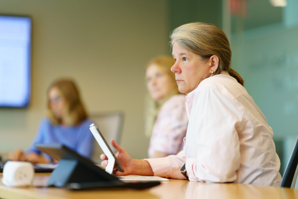 Gina Vanderloop looks up from her tablet toward the discussion, teammates blurred across the conference table.