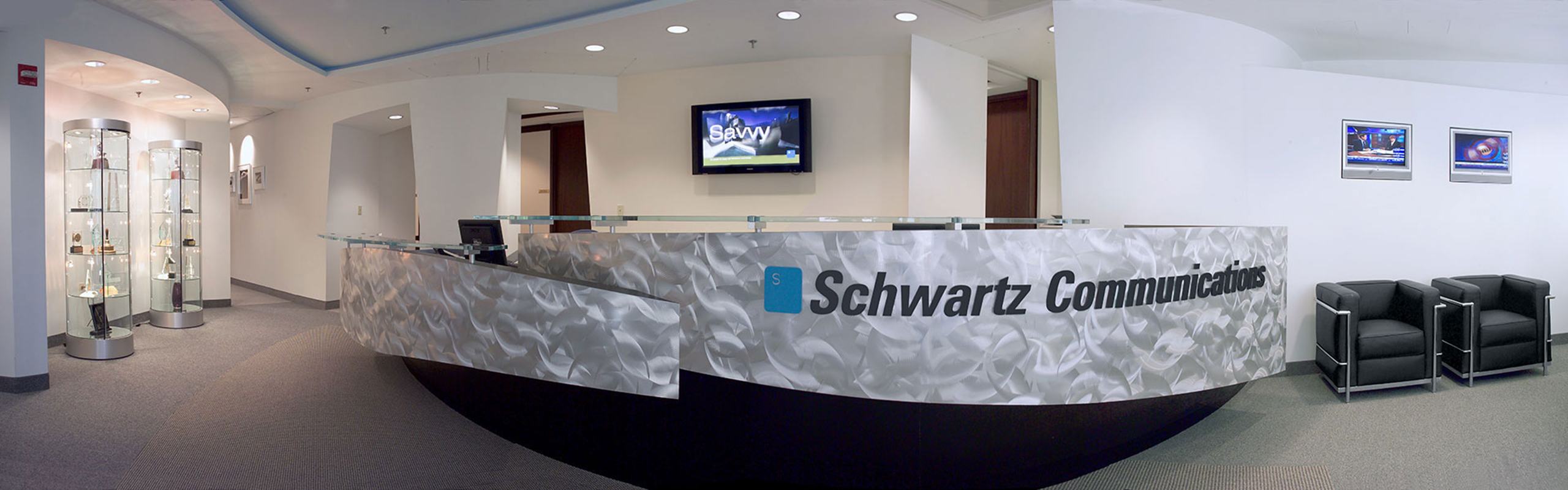 Panoramic reception lobby with the logo across a brushed-steel curved desk, display cases and leather chairs flanking the space.