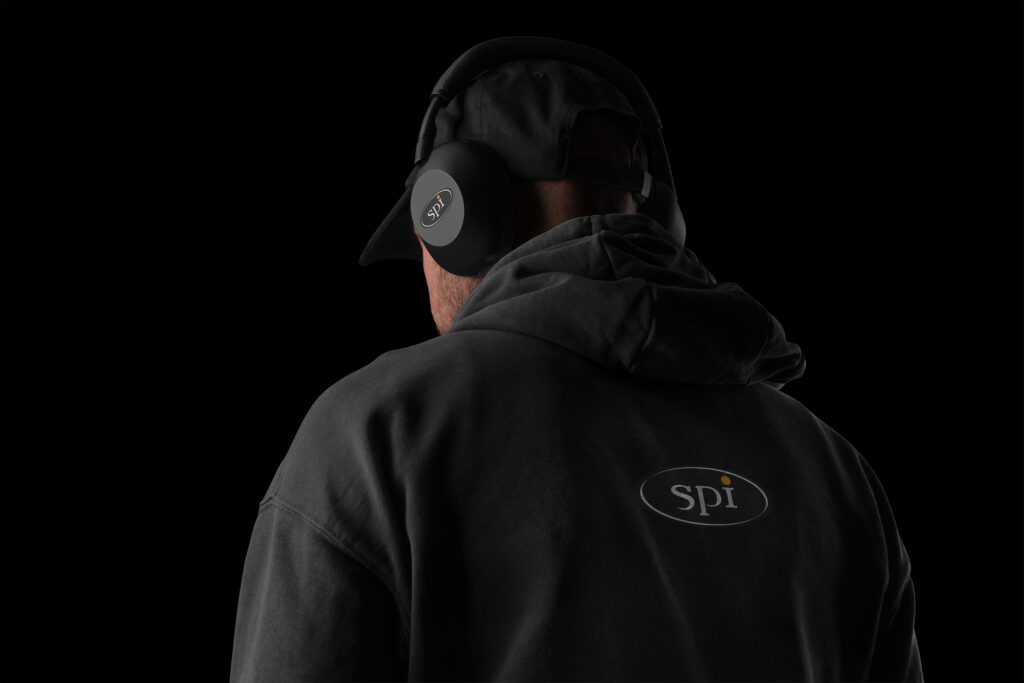 Hooded figure from behind in blackout tones, the SPI oval printed across the hoodie’s back echoing the badge on the headphone cup, the orange-dotted i the only color.