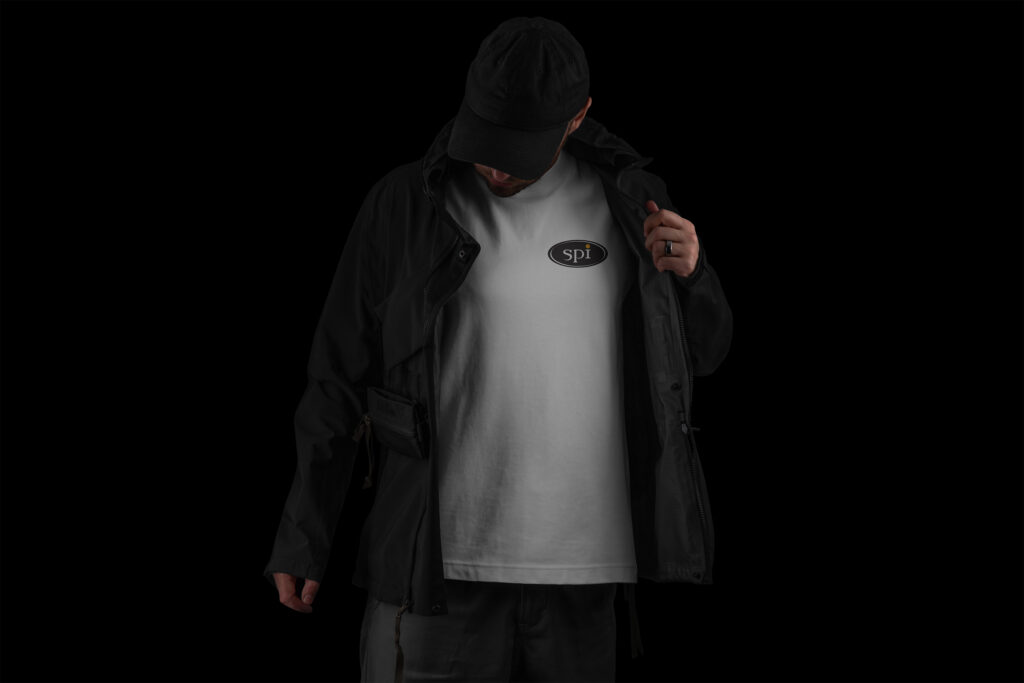 Gray SPI tee under an open black jacket and cap, the oval chest logo centered in the shadowed studio shot.