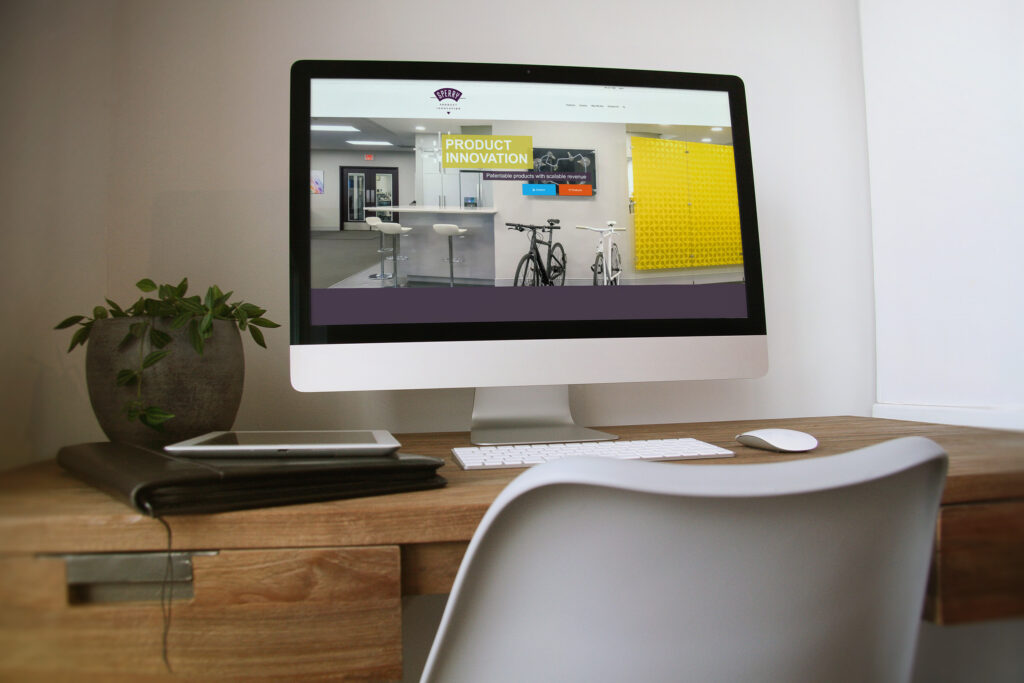 SPI homepage on an iMac, the Product Innovation banner over the workshop panorama of bikes and the yellow pegboard wall.