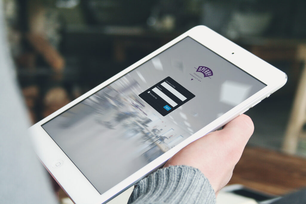 Partner login on an iPad mini, the SPI badge above credential fields against a blurred workshop, the doorway to shared files.
