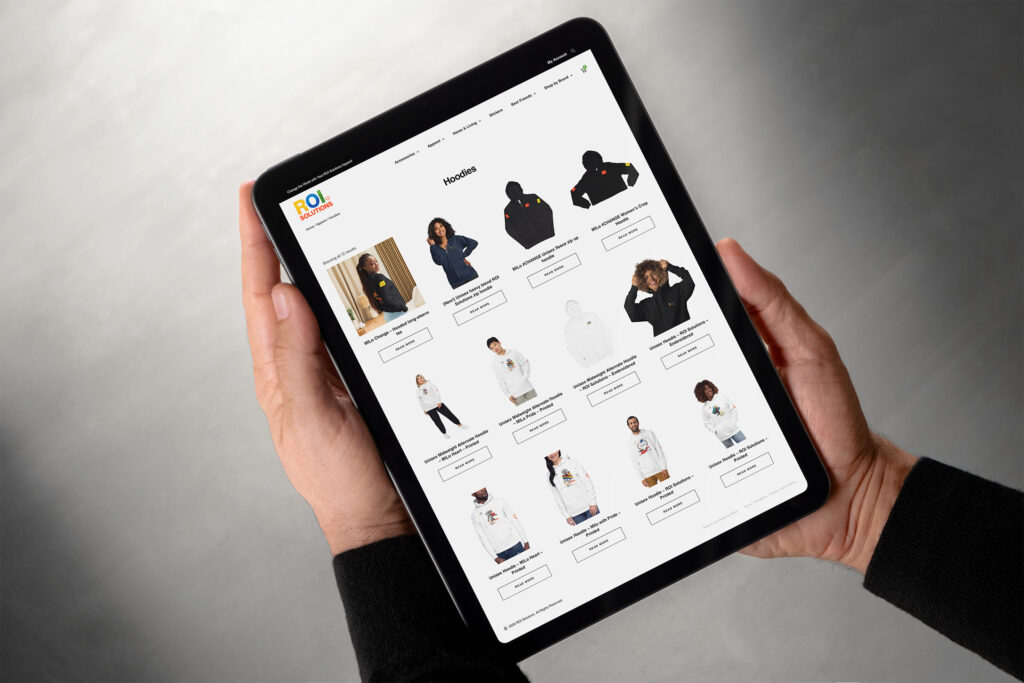 Hands hold a tablet showing the merch store hoodies category, product cards across a white grid.