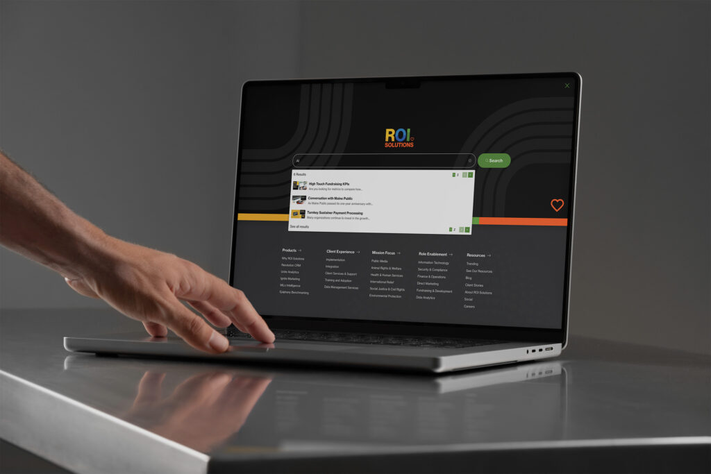 Hand at a laptop showing the dark predictive search overlay, results listed above the full navigation columns.