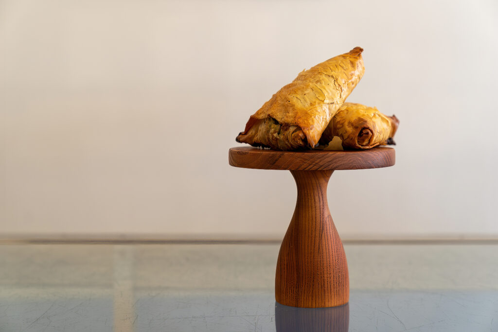 Two golden phyllo spanakopita triangles on a wooden pedestal at Arlington Bakery.