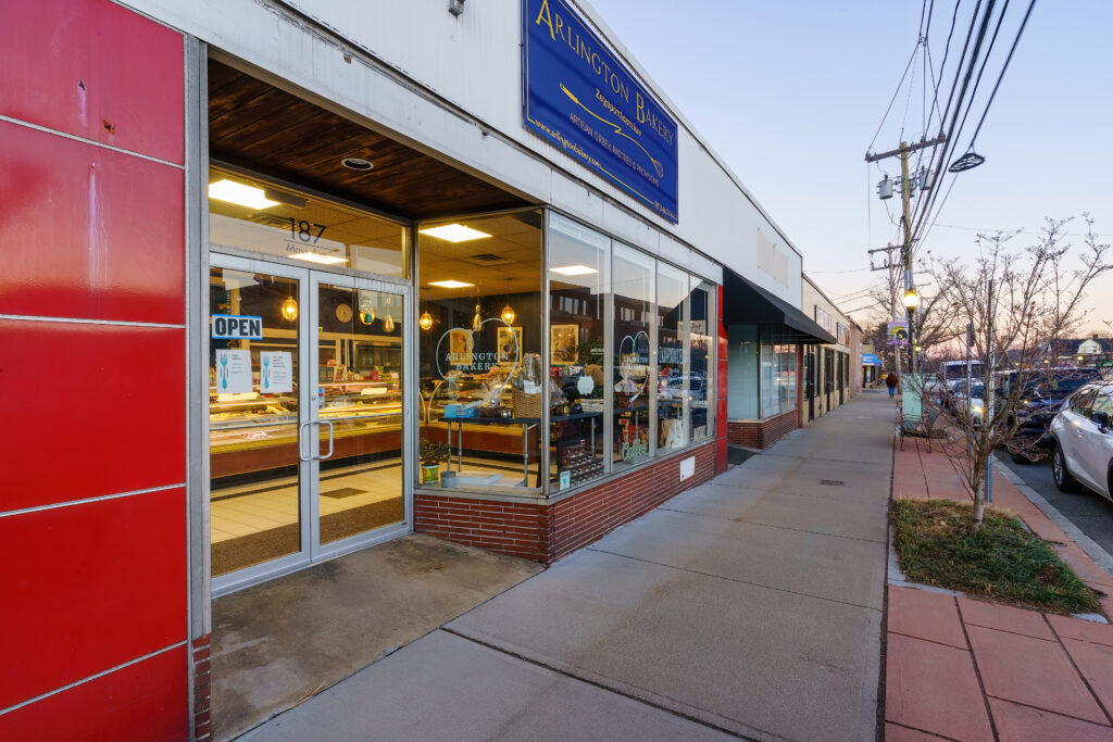 Arlington Bakery storefront on Massachusetts Avenue at dusk, red tile facade, OPEN sign lit and pastry cases visible through the glass.