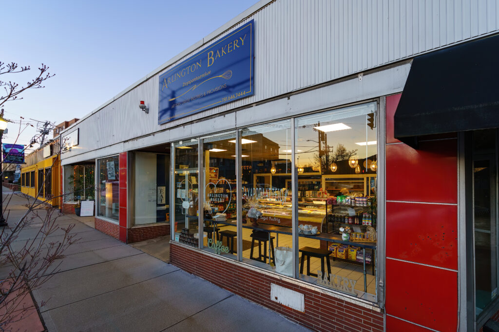 Arlington Bakery seen along the sidewalk at dusk, blue sign with rolling pin logo above red and white facade.