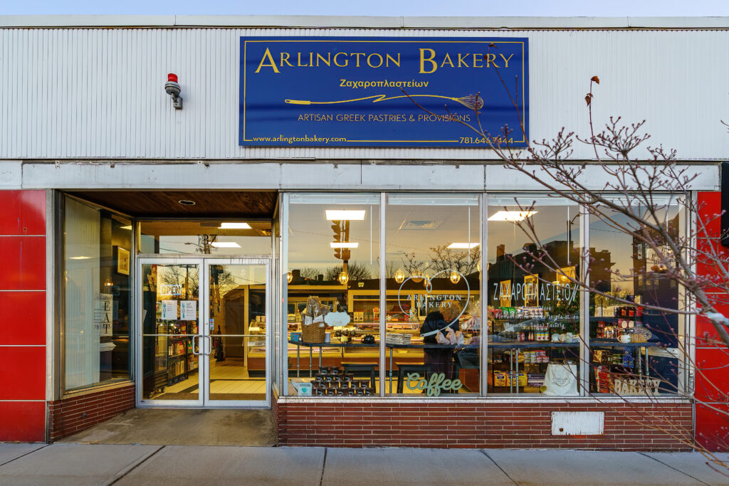 Front of Arlington Bakery with its blue Artisan Greek Pastries and Provisions sign, lit display windows at twilight.