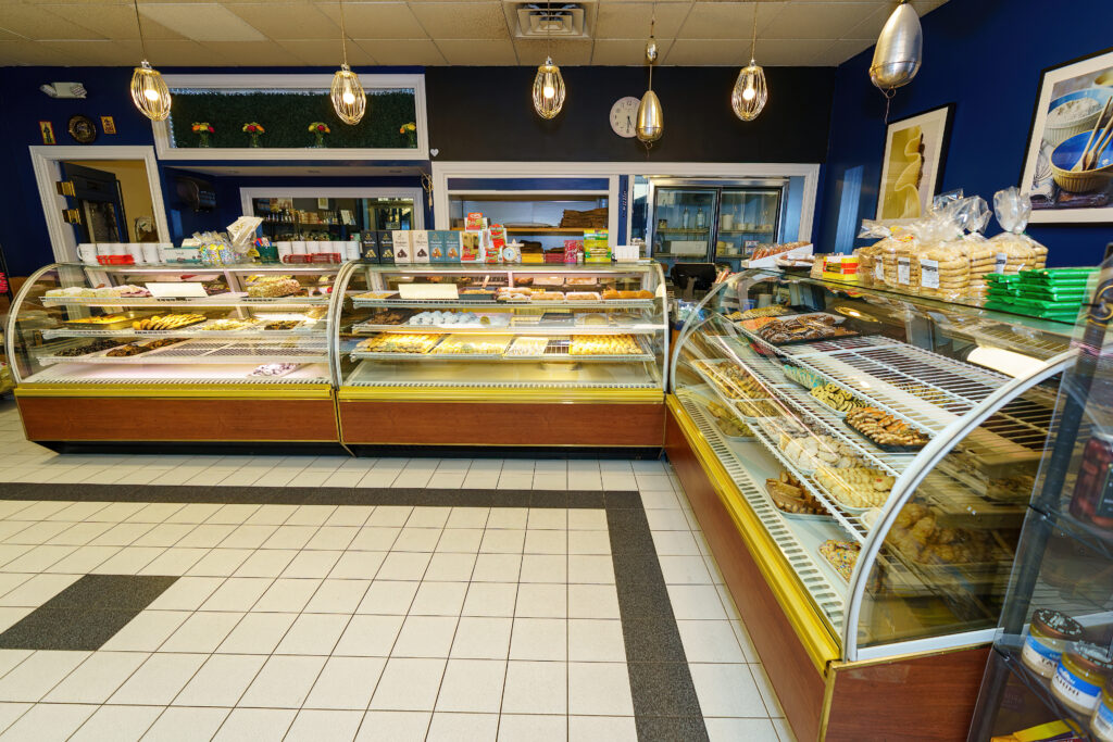 Arlington Bakery counter with pastry and cake cases full of Greek sweets, pendant lights overhead.