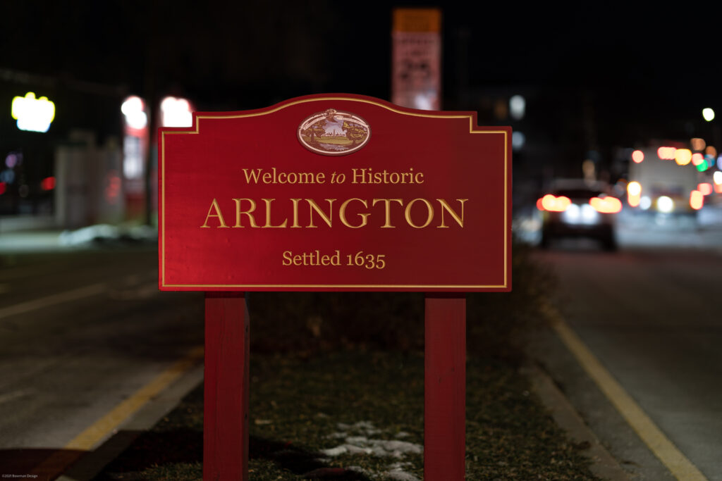 Red Welcome to Historic Arlington, Settled 1635 sign at night with headlights and traffic behind it.