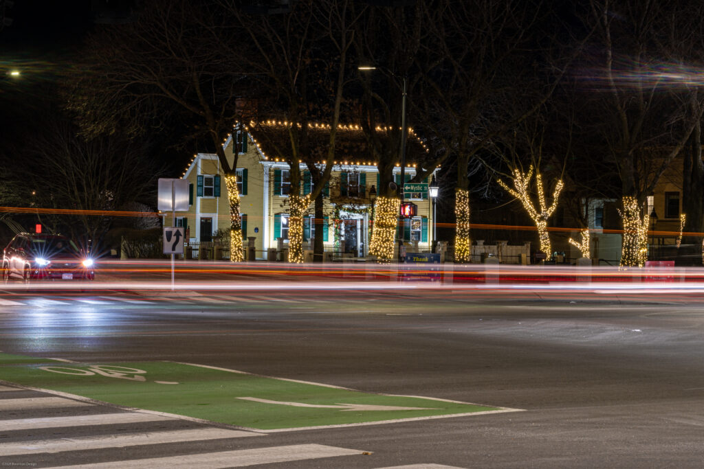 Long-exposure car light trails across a crosswalk in Arlington Center with lit trees and Cyrus Dallin Art Museum beyond.