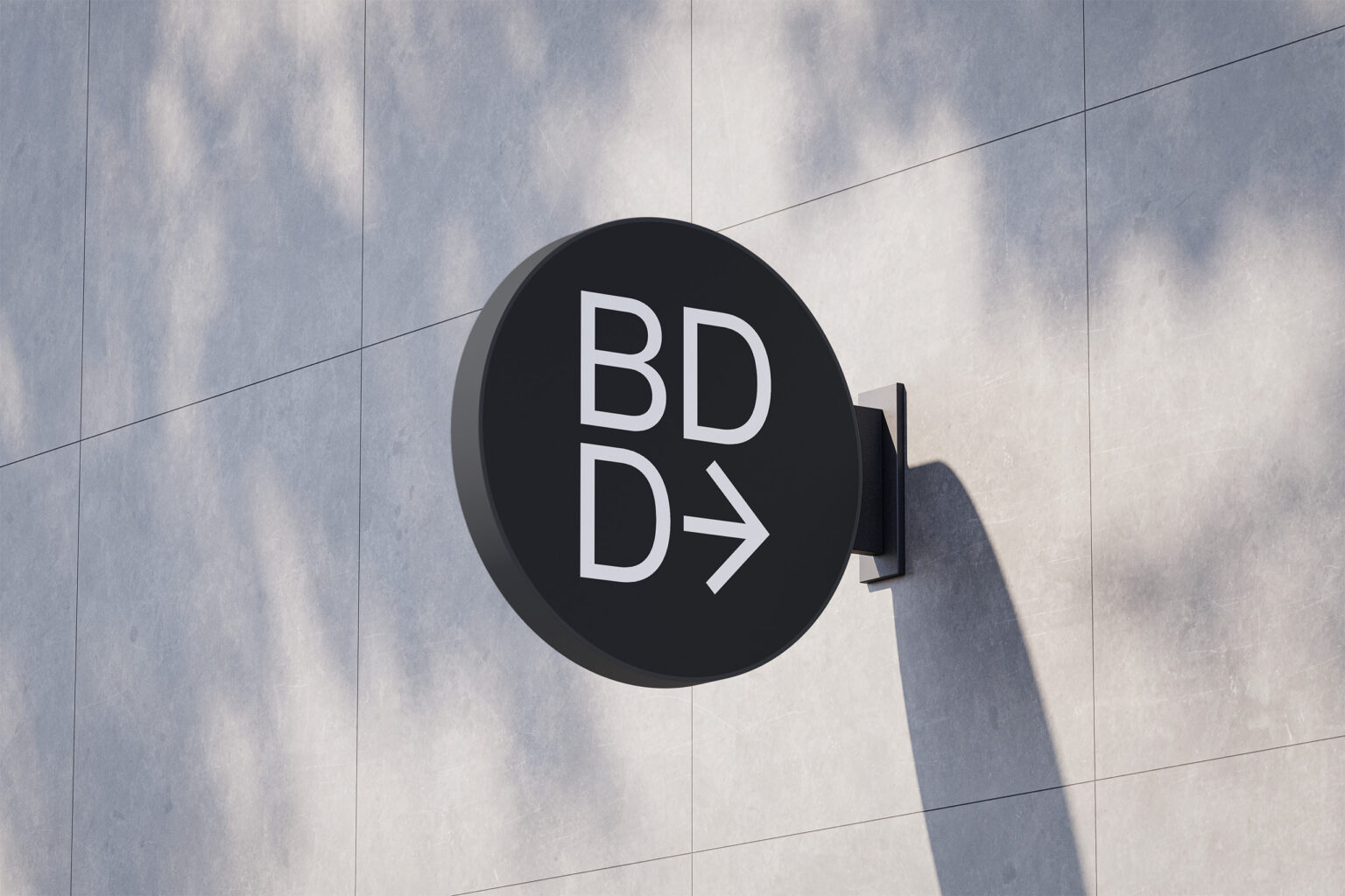 Black circular blade sign carrying the white BDD monogram and arrow, mounted to a sunlit gray stone facade with dappled tree shadows across the wall.