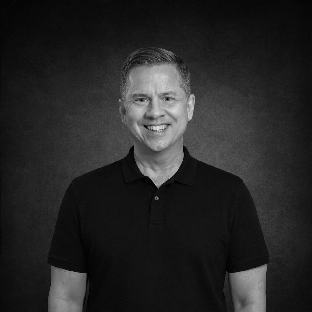 Black and white studio portrait of Bill Bowman, founder of Bowman Design Direction, smiling in a black polo shirt against a dark textured backdrop.