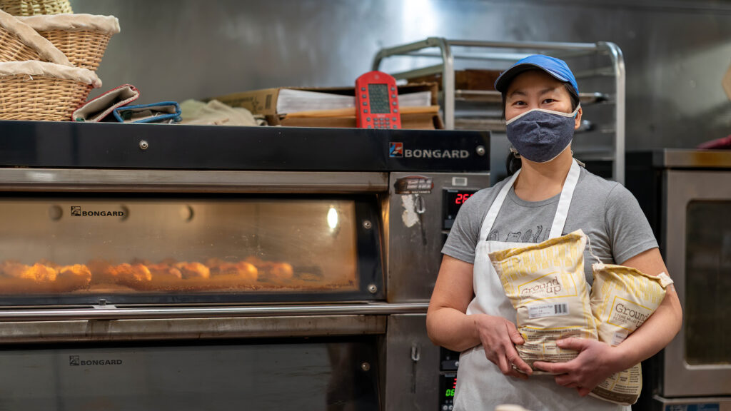 Baker Daisy Chow in a blue cap and mask holding two sacks of flour beside the Bongard deck oven at Breadboard Bakery.