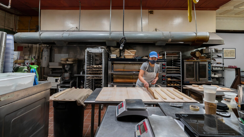Baker rolling long baguette dough on wooden boards in the Breadboard Bakery kitchen, deck oven behind.