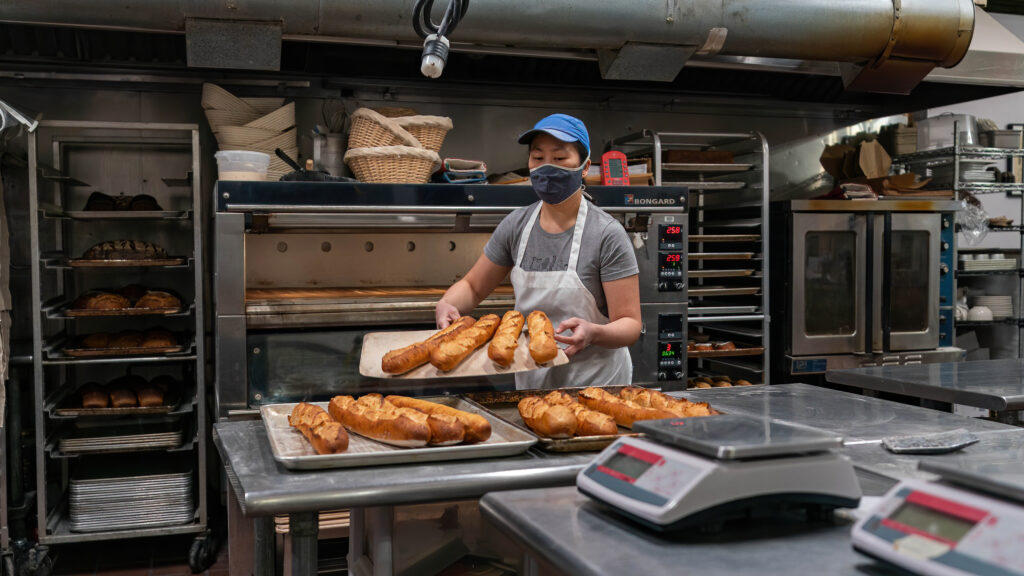 Daisy Chow carrying a sheet tray of golden baguettes across the Breadboard Bakery kitchen.