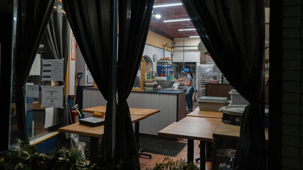 View through Breadboard Bakery's front window past dark curtains to the counter and baker at work.