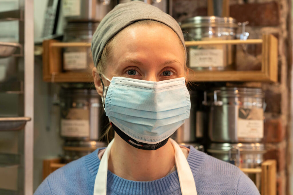 Butternut Bakehouse team member in a gray knit hat and mask in front of shelves of jarred goods.