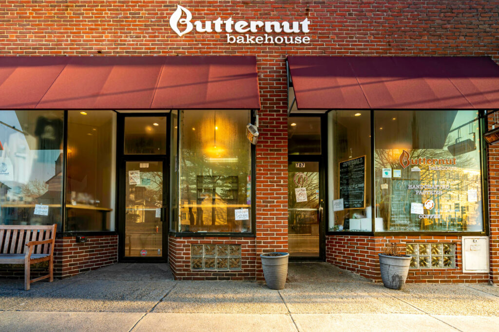 Front of Butternut Bakehouse, brick facade with two red awnings and a wooden bench, Arlington MA.