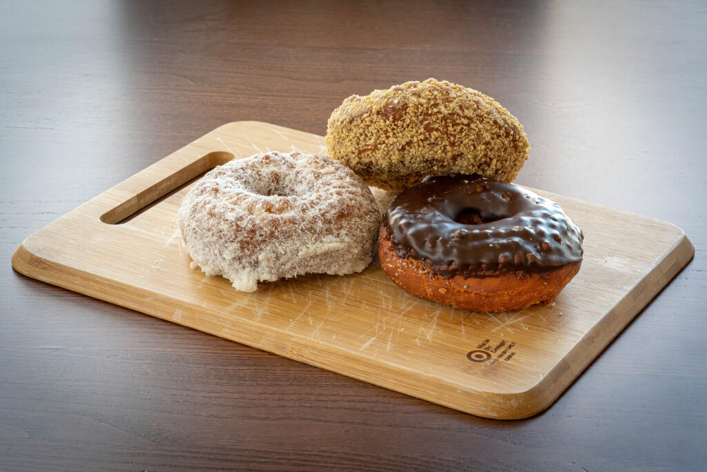 Three Gail Ann donuts, powdered, crumb and chocolate frosted, on a wooden board.