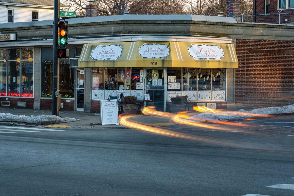 Magic Bites Bakery and Cafe corner storefront with car light trails passing at dusk, Arlington MA.