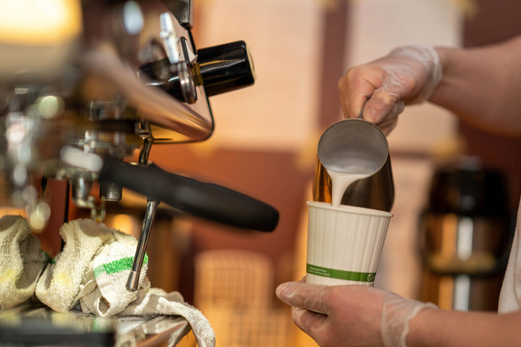 Barista pouring steamed milk from a pitcher into a paper cup at The Roasted Granola.