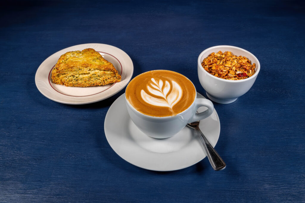 Latte with leaf art, a scone and a bowl of granola on a blue table at The Roasted Granola.