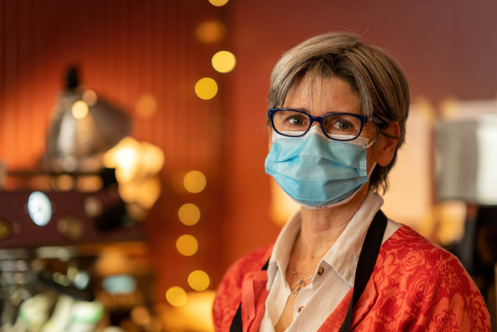 Owner of The Roasted Granola in glasses and a mask, red patterned top, with string lights behind.