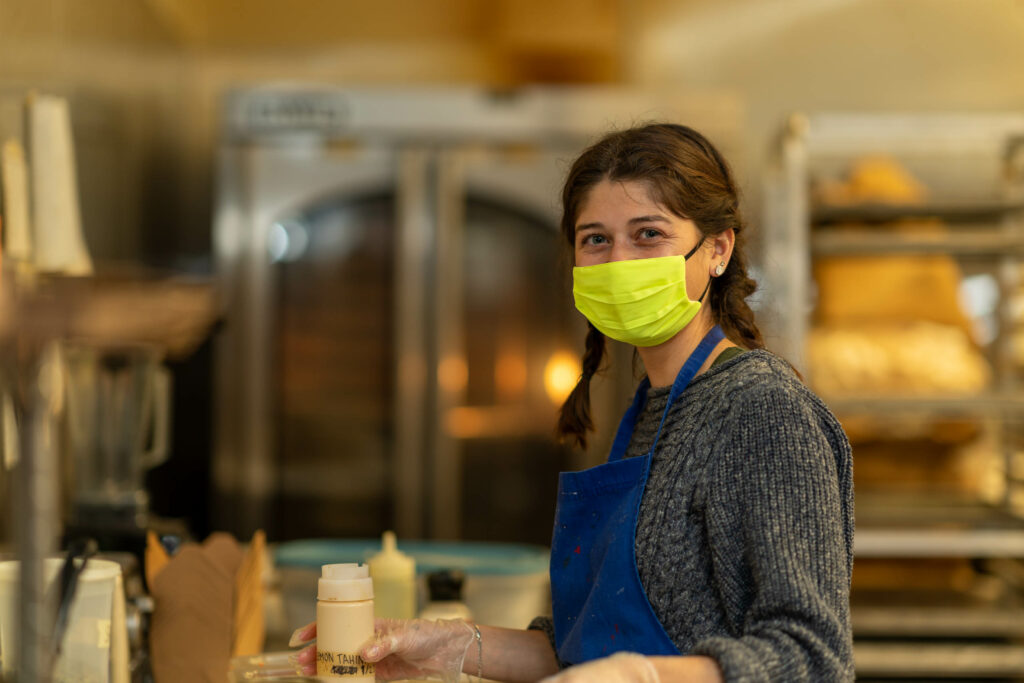 Team member in a yellow mask and blue apron working at the kitchen counter, The Roasted Granola.