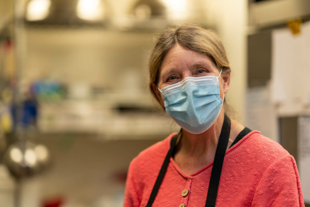 Team member in a coral top, black apron and blue mask in The Roasted Granola kitchen.