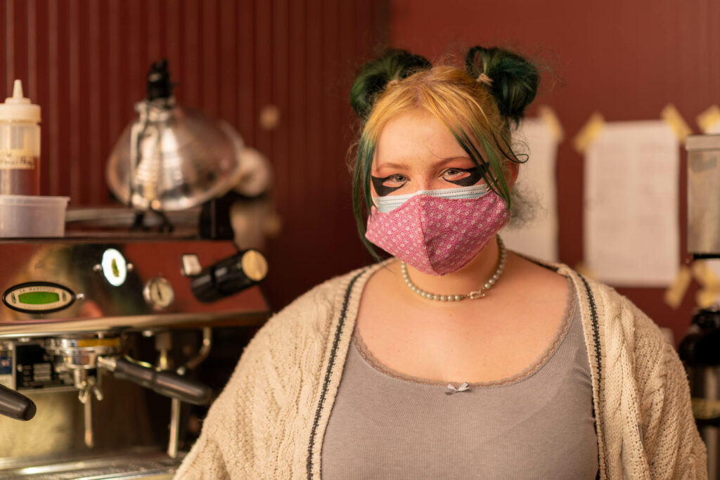 Barista with green and blonde space buns in a pink mask in front of the espresso machine at The Roasted Granola.