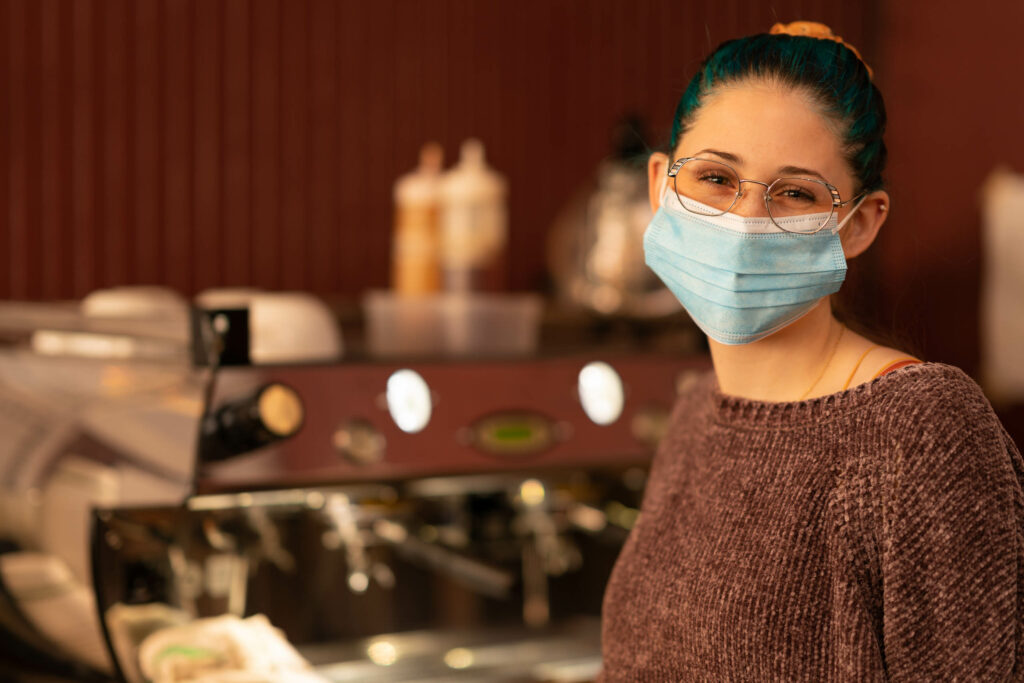Barista in glasses and a blue mask beside the espresso machine at The Roasted Granola.