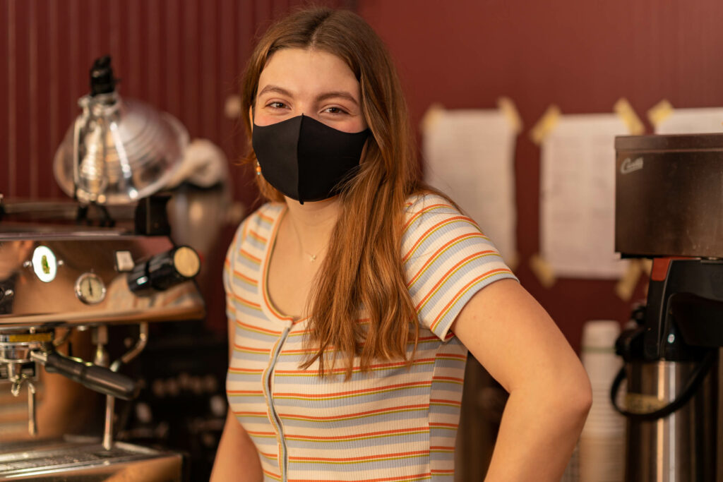 Barista in a striped shirt and black mask beside the espresso machine at The Roasted Granola.