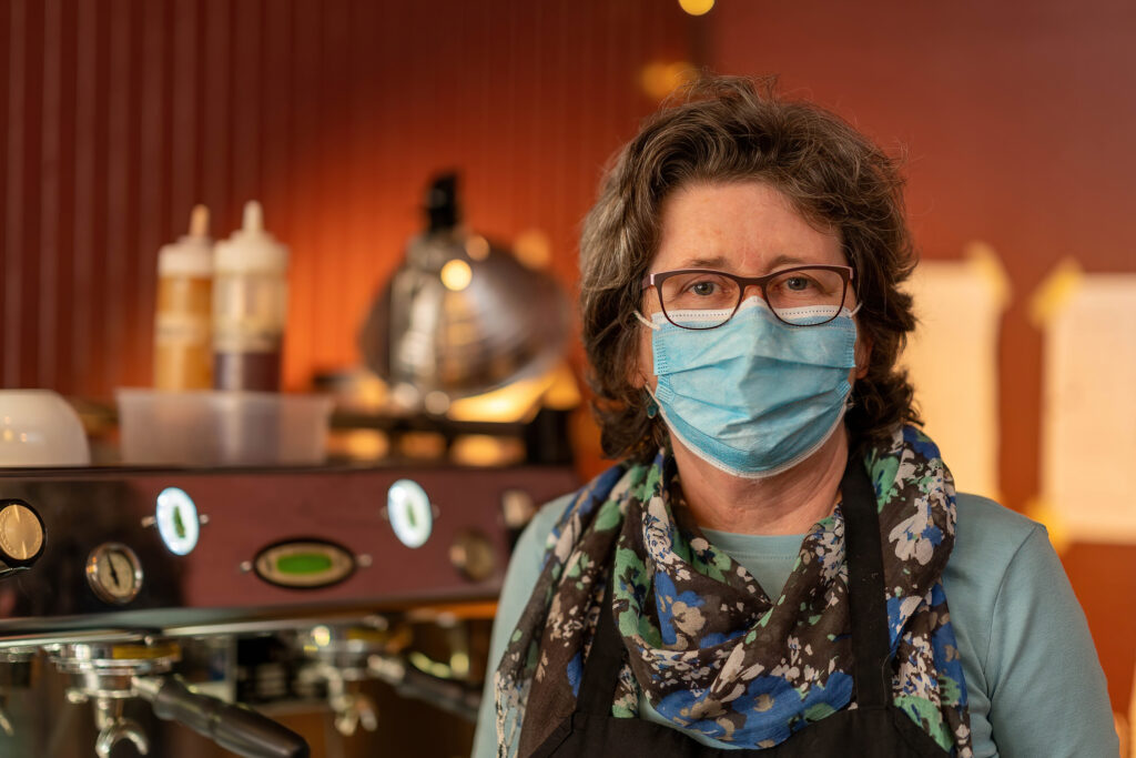 Owner of The Roasted Granola in a floral scarf and apron standing beside the espresso machine.